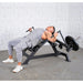 A man in a grey athletic outfit performing a weight leverage pec fly bench press on a black adjustable weight bench in a gym setting with a textured white wall and concrete floor.