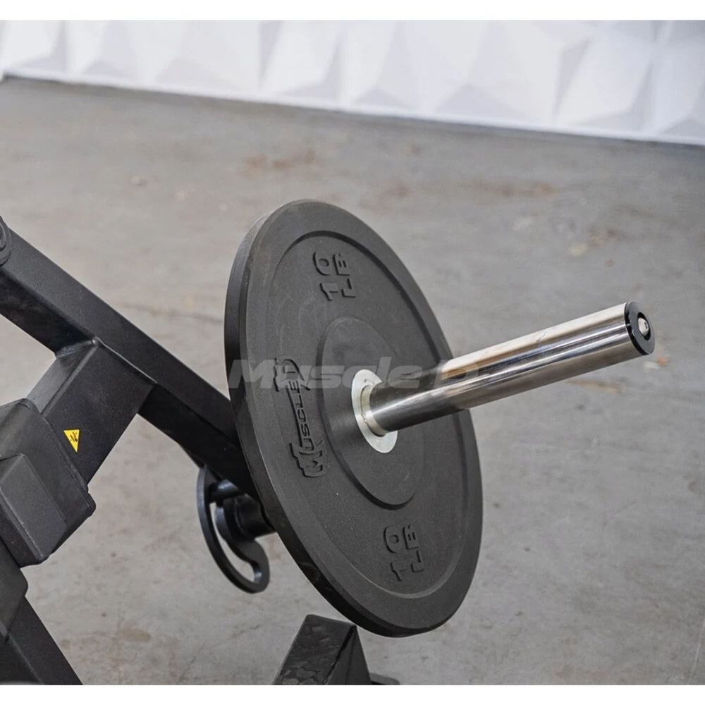 A close-up of a black weight plate on a barbell mounted on a black exercise machine, with a concrete floor and white draped backdrop in the background.