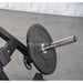 A close-up of a black weight plate on a barbell mounted on a black exercise machine, with a concrete floor and white draped backdrop in the background.