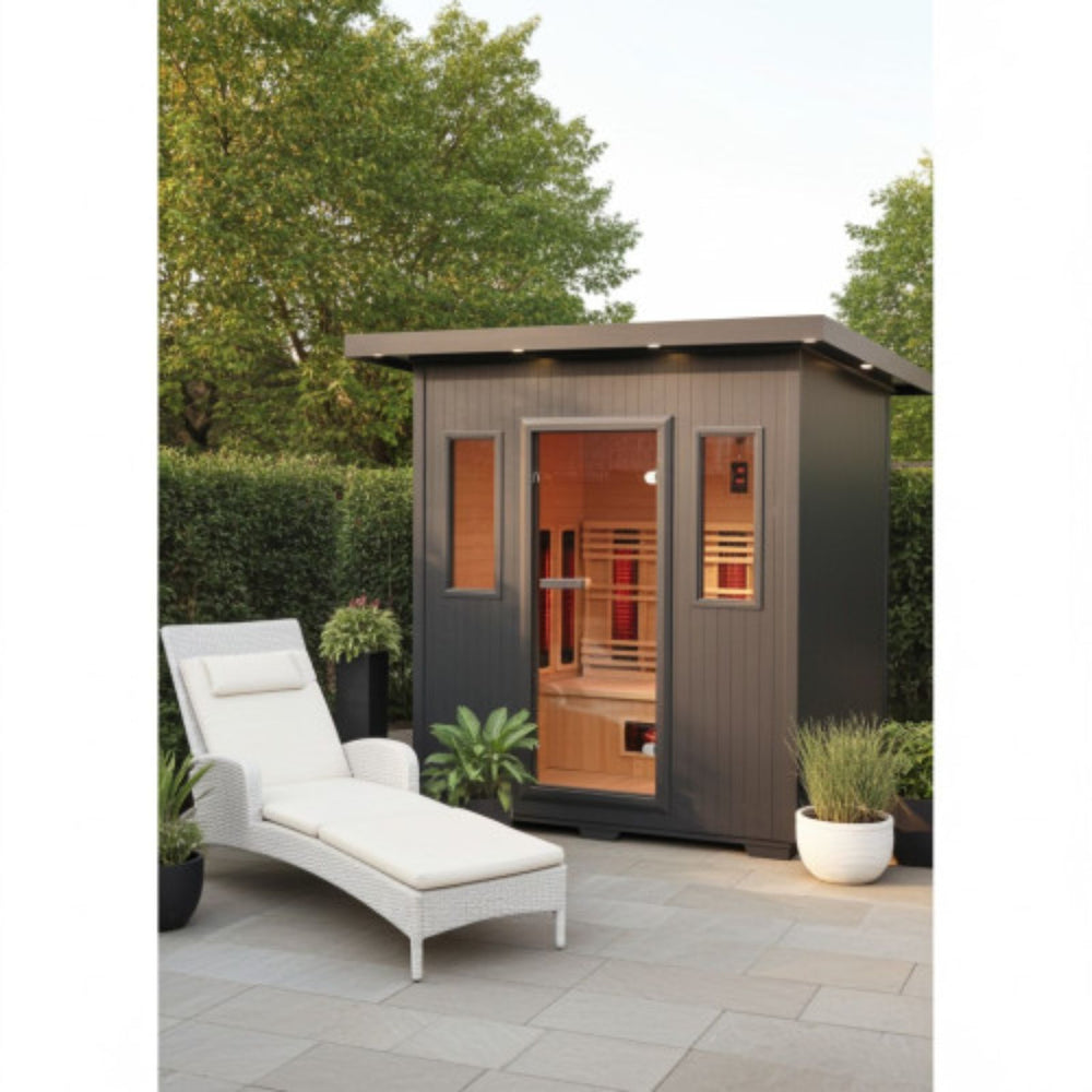 A dark gray outdoor Sunray Pacific 4-Person Outdoor Infrared Sauna cabin with a glass door and side windows, shown in a landscaped patio setting with a white lounge chair, potted plants, and trees.