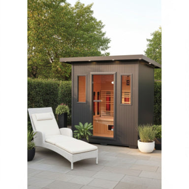 A dark gray outdoor Sunray Pacific 4-Person Outdoor Infrared Sauna cabin with a glass door and side windows, shown in a landscaped patio setting with a white lounge chair, potted plants, and trees.