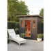 A dark gray outdoor Sunray Pacific 4-Person Outdoor Infrared Sauna cabin with a glass door and side windows, shown in a landscaped patio setting with a white lounge chair, potted plants, and trees.