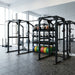 A black TKO Annex Dual Power Rack & Storage System 920PR-ANX multi-functional power rack in a modern gym setting, featuring a built-in storage shelf with kettlebells and colorful weight plates, with other gym equipment visible in the background.